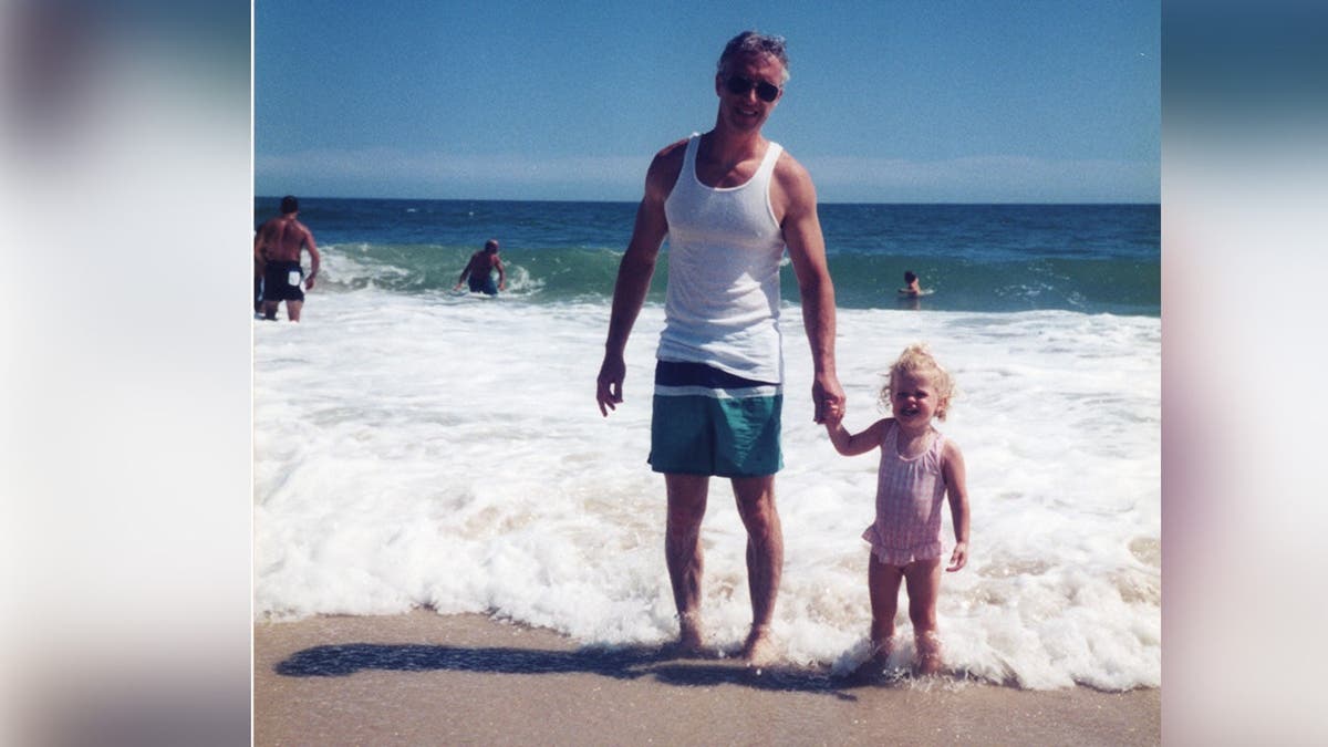 image shows father holding little girls hand at the beach