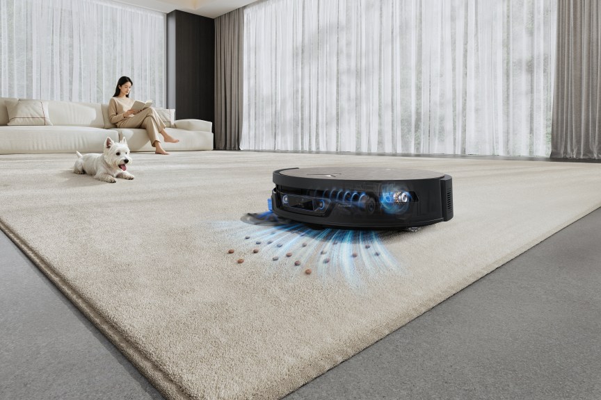 Robovac picks up debris from a carpet as a person sits on a couch and a pet dog sits nearby.